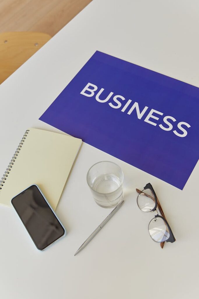 Flat lay of a desk with business-themed poster, notebook, smartphone, pen, eyeglasses, and water glass.